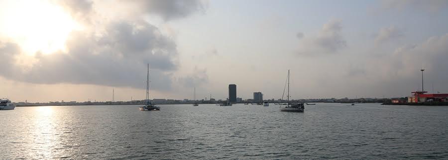 calm grey water and a cloudy sky looking into the sun with a few yachts at anchor and a container port to the right