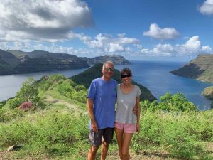 the couple who this article are about are dressed in shorts and tshirts with sunglasses standing on a headland very green and fertile with a bay in the background