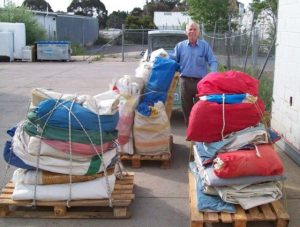 pallets stacked high with brightly colored sails tied up with rope and a man in a blue shirt and brown trousers standing amongst them