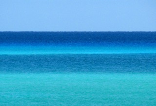 horizontal lines of bright blue sea of varying shades topped by a pale blue solid colour sky not a cloud in sight