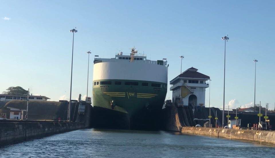 a huge ship in the canal almost touching the sides with a green hull and white topsides taller than the canal admin building