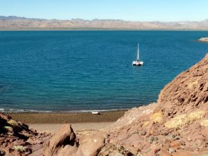 lone catamaran anchored in a bay off a brown volcanic beach with a very dry landscape all around